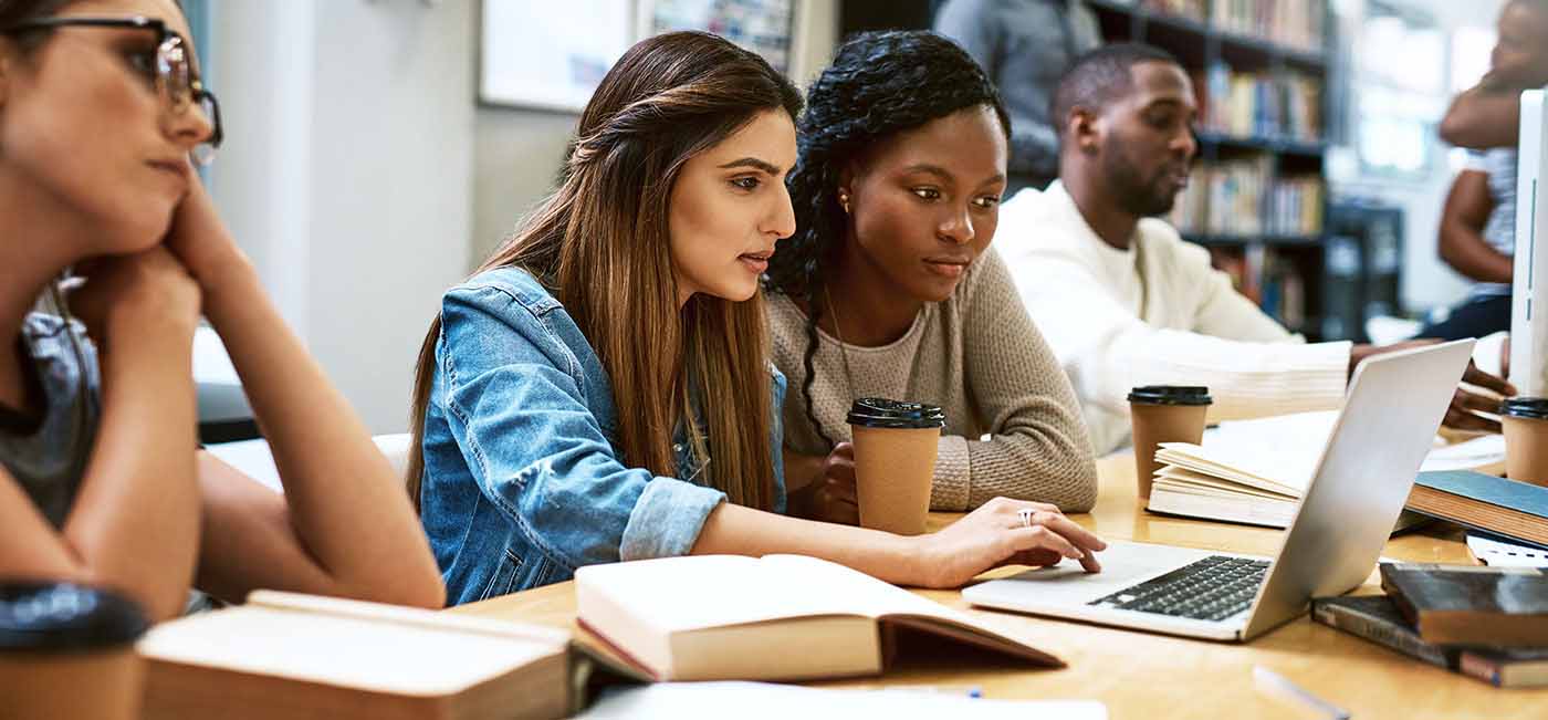 Four students sitting in library, studying on laptop with coffee